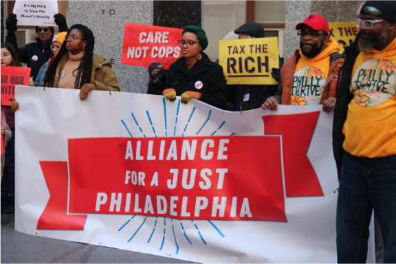 People outside City Hall holding signs and a banner saying Alliance for a Just Philadelphia