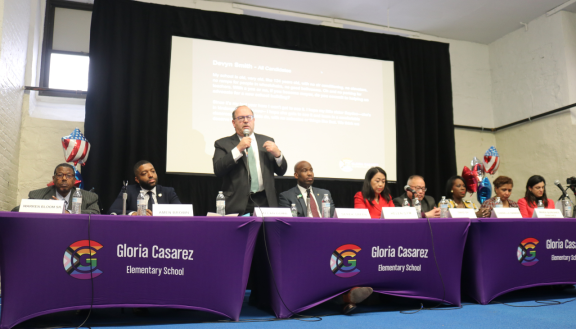 Candidates sit at tables in front of a large screen at Gloria Casarez Elementary School forum