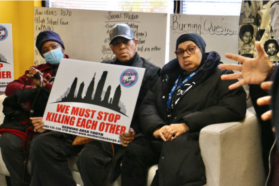 Three members of the anti-gun violence group Mothers In Charge sit on a couch with a sign saying We Must Stop Killing Each Other