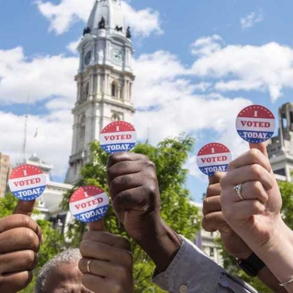 Seven hands with I voted stickers on their thumbs and Philadelphia City Hall in the background