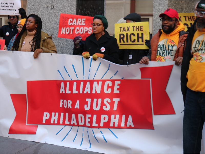 People outside City Hall holding signs and a banner saying Alliance for a Just Philadelphia