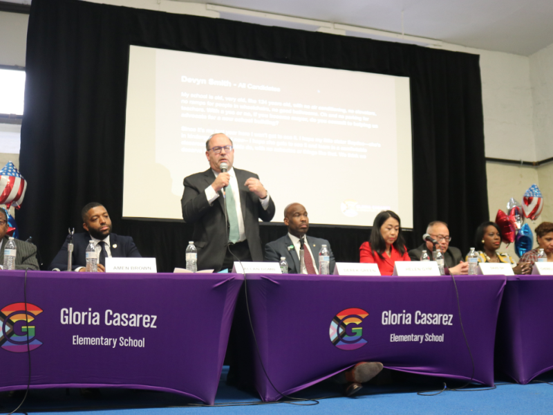 Candidates sit at tables in front of a large screen at Gloria Casarez Elementary School forum