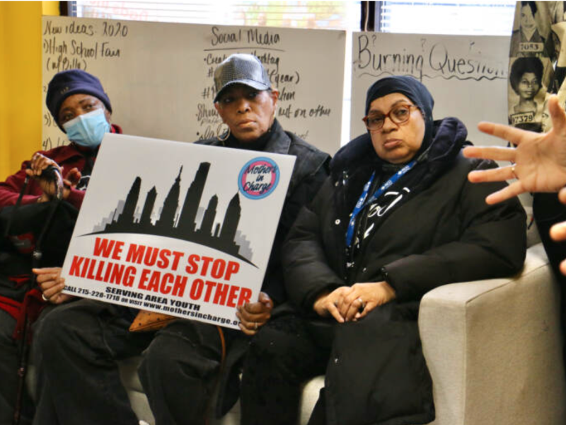 Three members of the anti-gun violence group Mothers In Charge sit on a couch with a sign saying We Must Stop Killing Each Other