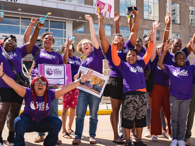 Students and adults in purple "Life Every Voice" shirts cheering in front of a school