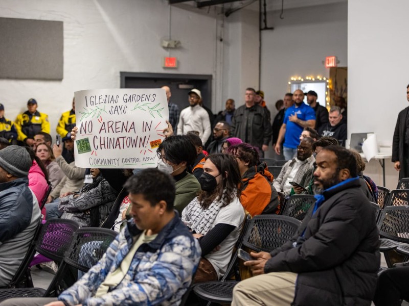 Community members listening to mayor at a town hall meeting. One community member holds a sign opposing an arena being built in Chinatown