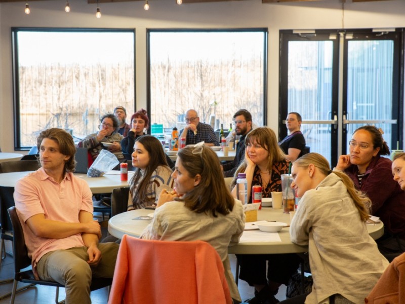 A diverse group of people sits around round tables in a bright, casual indoor setting, attentively listening to a presentation or speaker off-camera.
