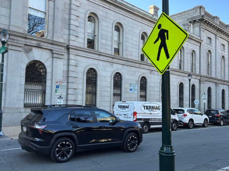 A bright yellow pedestrian crossing sign is posted along a city street lined with parked cars and a historic stone building, as a black SUV drives by.